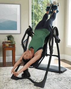 Woman using an inversion table for back stretching and relaxation in a bright, modern living room, featuring a large window with greenery outside and a stylish rug.