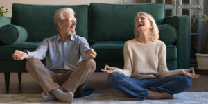 Elderly man and woman sitting on a living room floor, laughing and practicing mindfulness or meditation. They are dressed casually, with a green sofa in the background, creating a warm and inviting atmosphere.