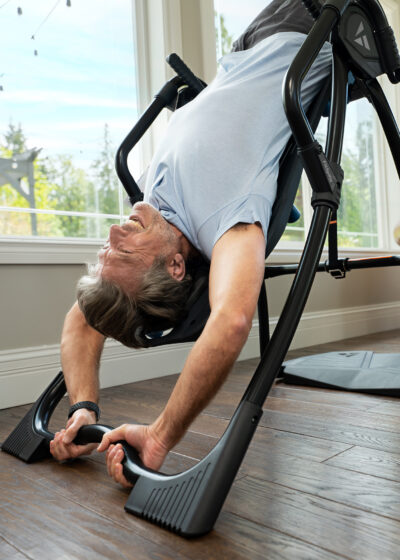 A man using an inversion table for back stretching and relaxation, positioned in a bright room with large windows and wooden flooring.